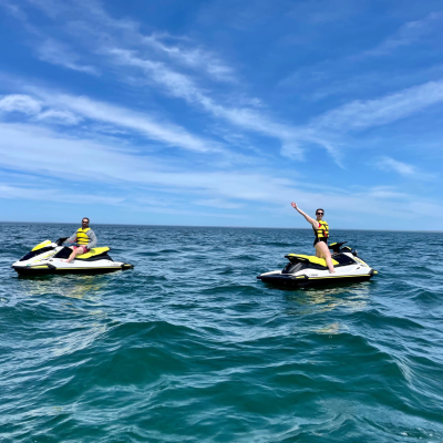 Couple Jetskiing Two guests riding jet skis on the open waters of Cape Cod Bay under a bright blue sky with scattered clouds. One rider waves while enjoying a guided jet ski adventure with Dennis Parasail & Jet Ski.
